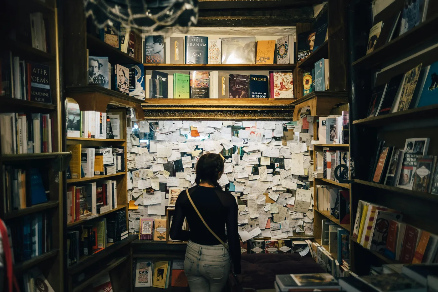 a nook in a book store with a frame full of notes, poems, or postcards and a women in front of them looking